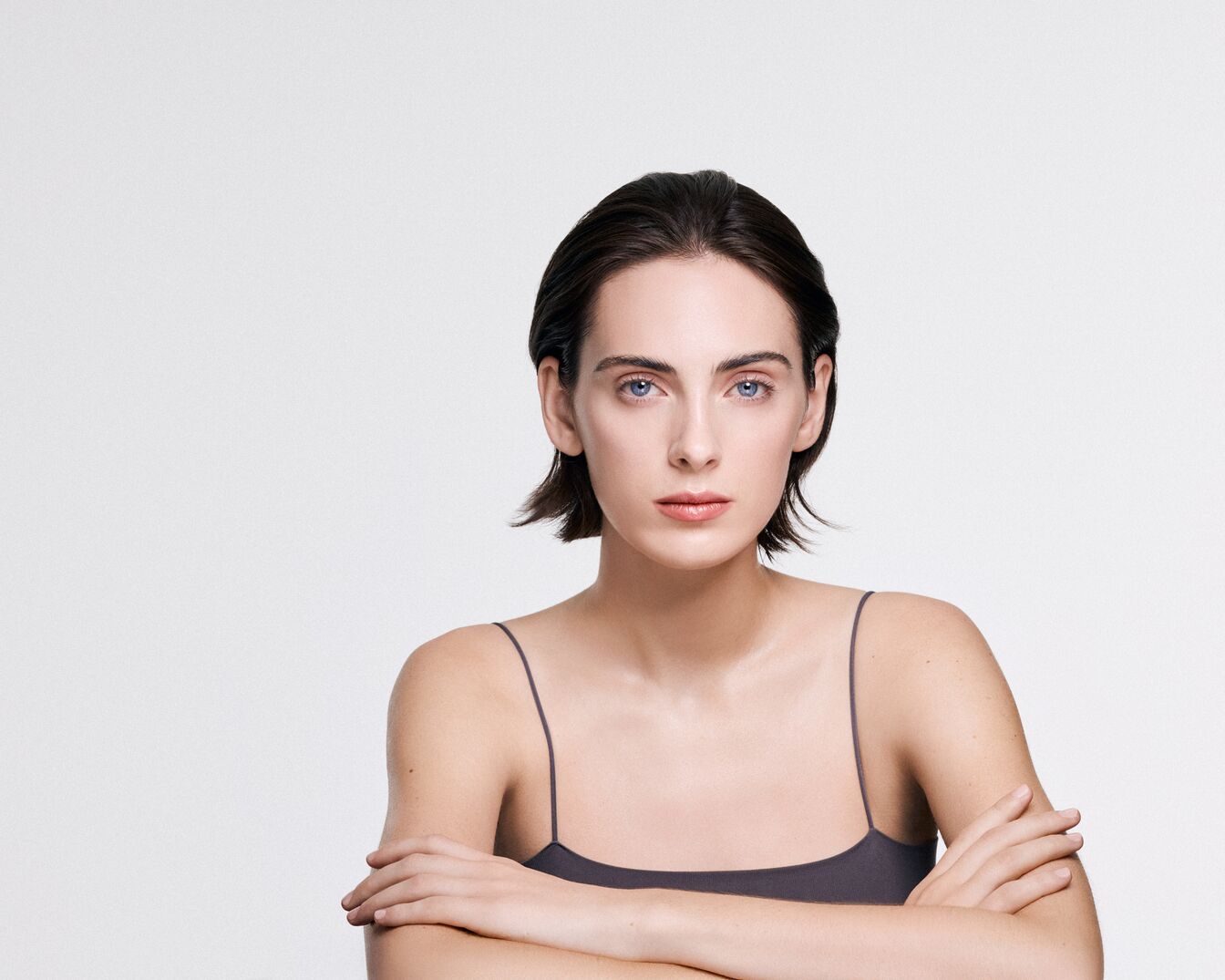 Una donna con capelli corti e scuri, occhi azzurri e labbra rosa chiaro, indossa una canotta grigio scuro con spalline sottili e ha le braccia incrociate.