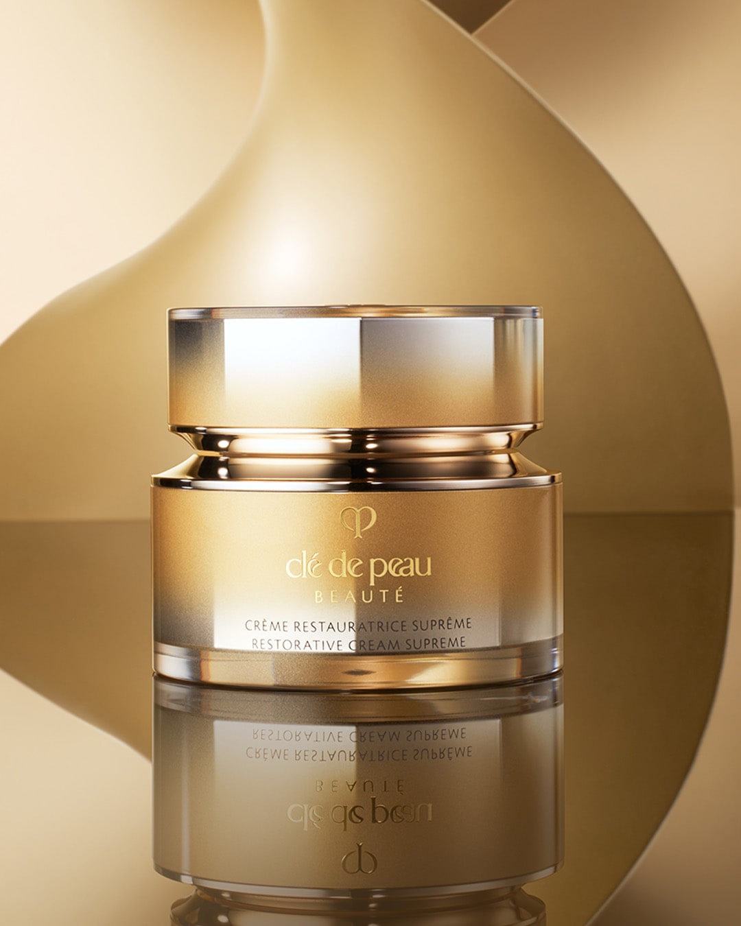 Die Clé de Peau Beauté Crème Restauratrice Suprême ist in einem eleganten, goldfarbenen Tiegel mit einem glänzenden Deckel verpackt und steht auf einer spiegelnden Oberfläche vor einem goldenen, geschwungenen Hintergrund.