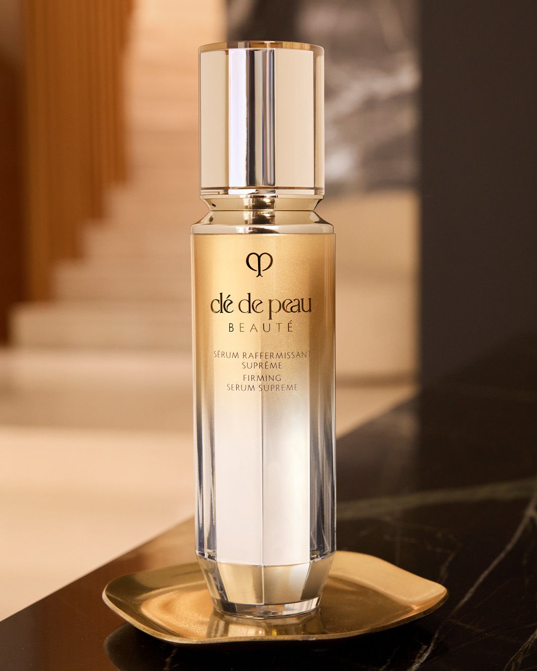 Das Clé de Peau Beauté Serum Supreme in einem eleganten, gold-silbernen Flakon steht auf einem goldenen Tablett und verspricht straffere Haut.