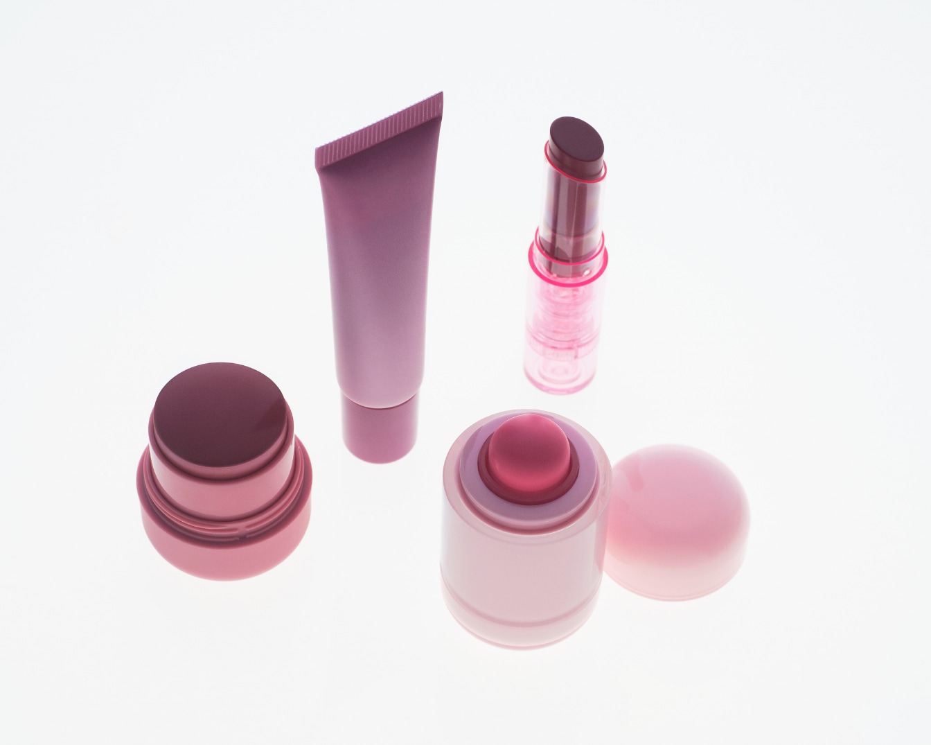 Un assortiment de produits de maquillage roses et violets, comprenant un tube de crème, un rouge à lèvres et deux baumes à lèvres, est présenté sur un fond blanc éclatant.