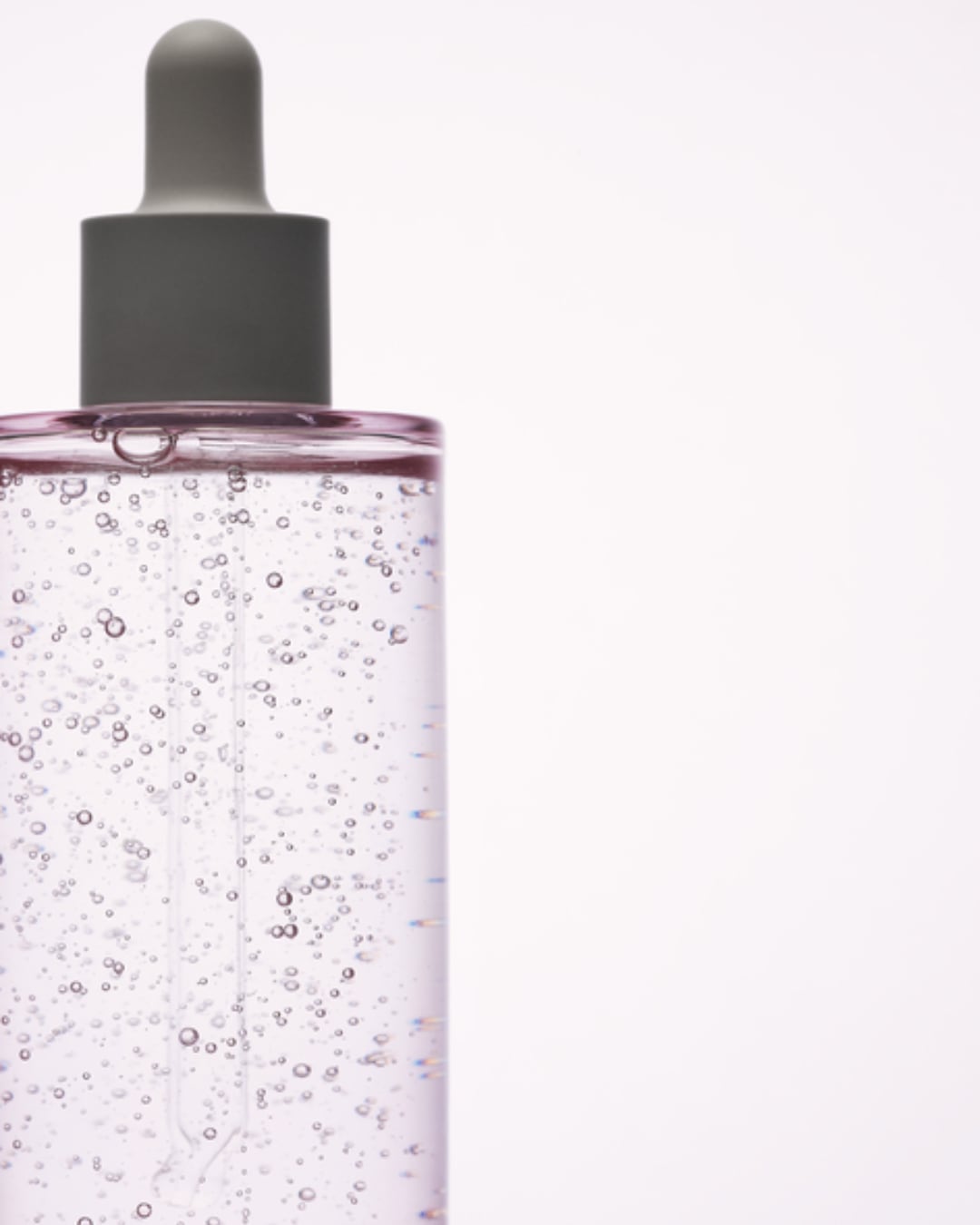 Un flacon de sérum transparent avec un compte-gouttes gris, rempli d'un liquide rose clair parsemé de bulles, sur un fond blanc.