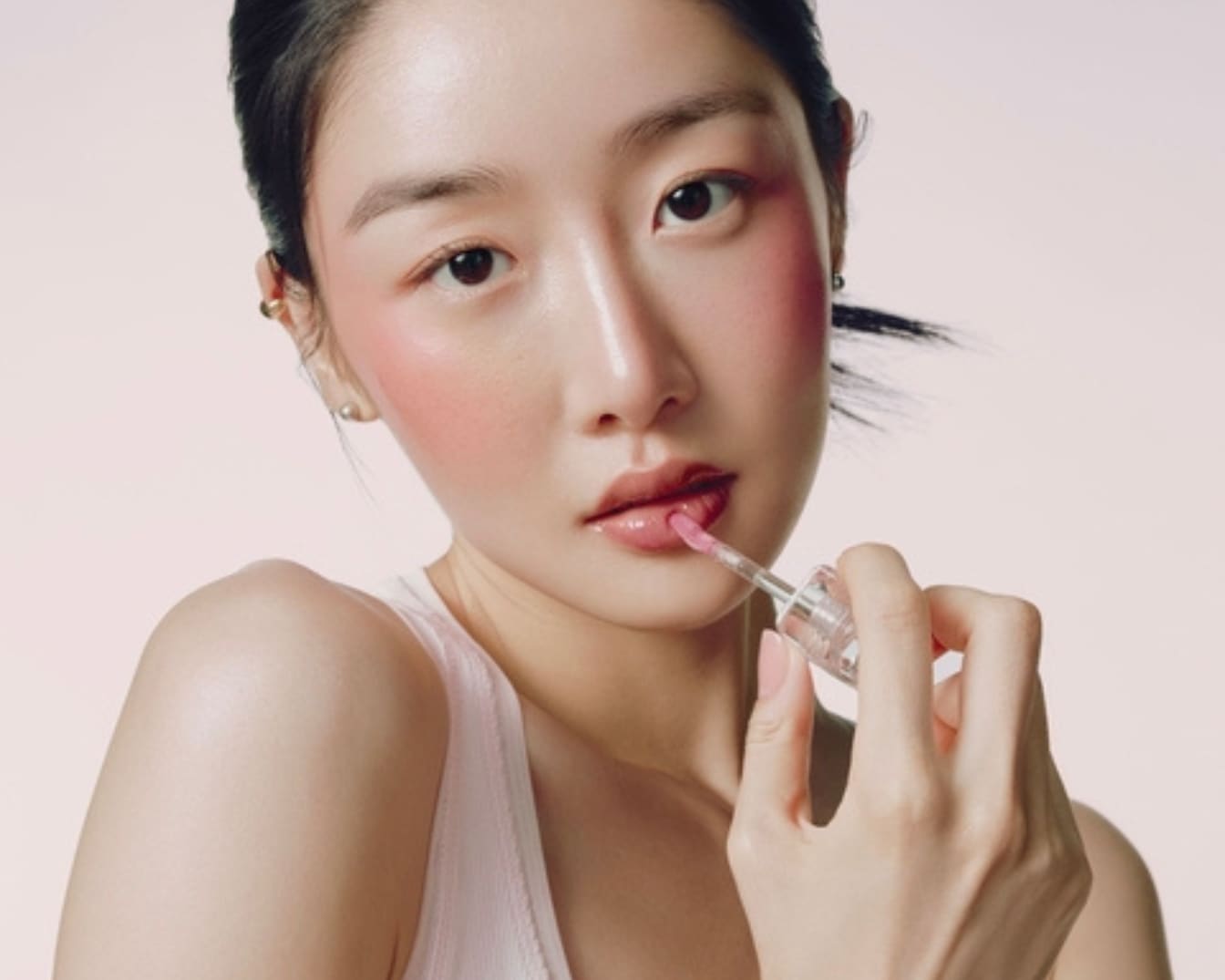 Une femme applique un brillant à lèvres rose clair sur ses lèvres, avec un fard à joues rose vif sur ses joues, créant un look frais et éclatant.