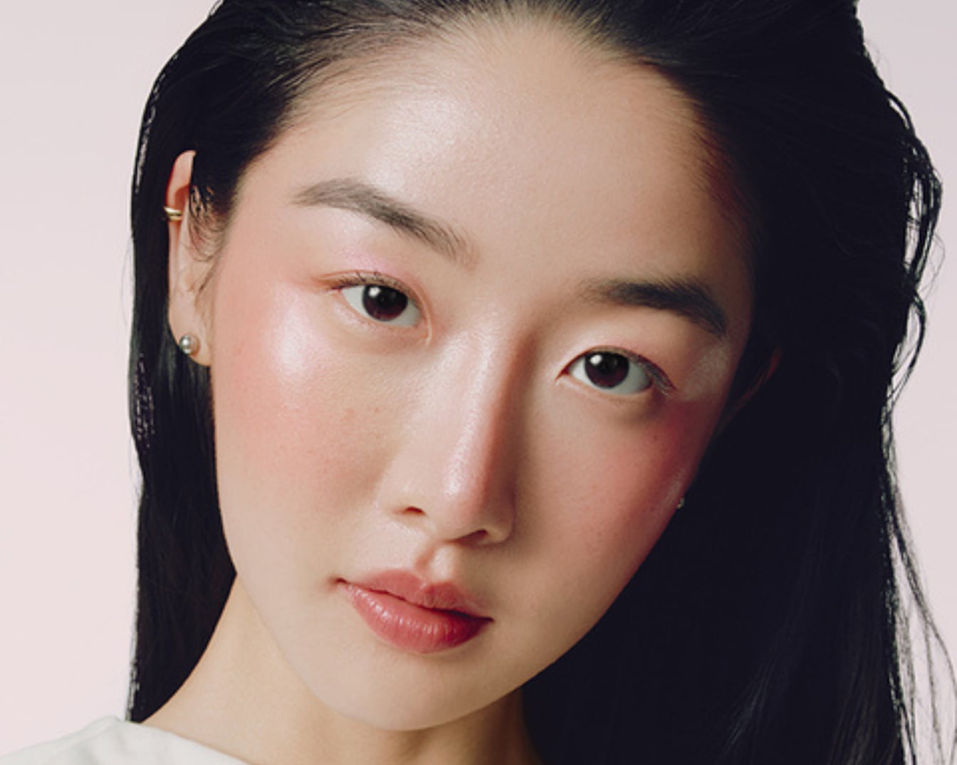 Une femme aux cheveux noirs et brillants, le visage maquillé de fard à joues rose et de fard à paupières rose, regarde directement l'objectif.