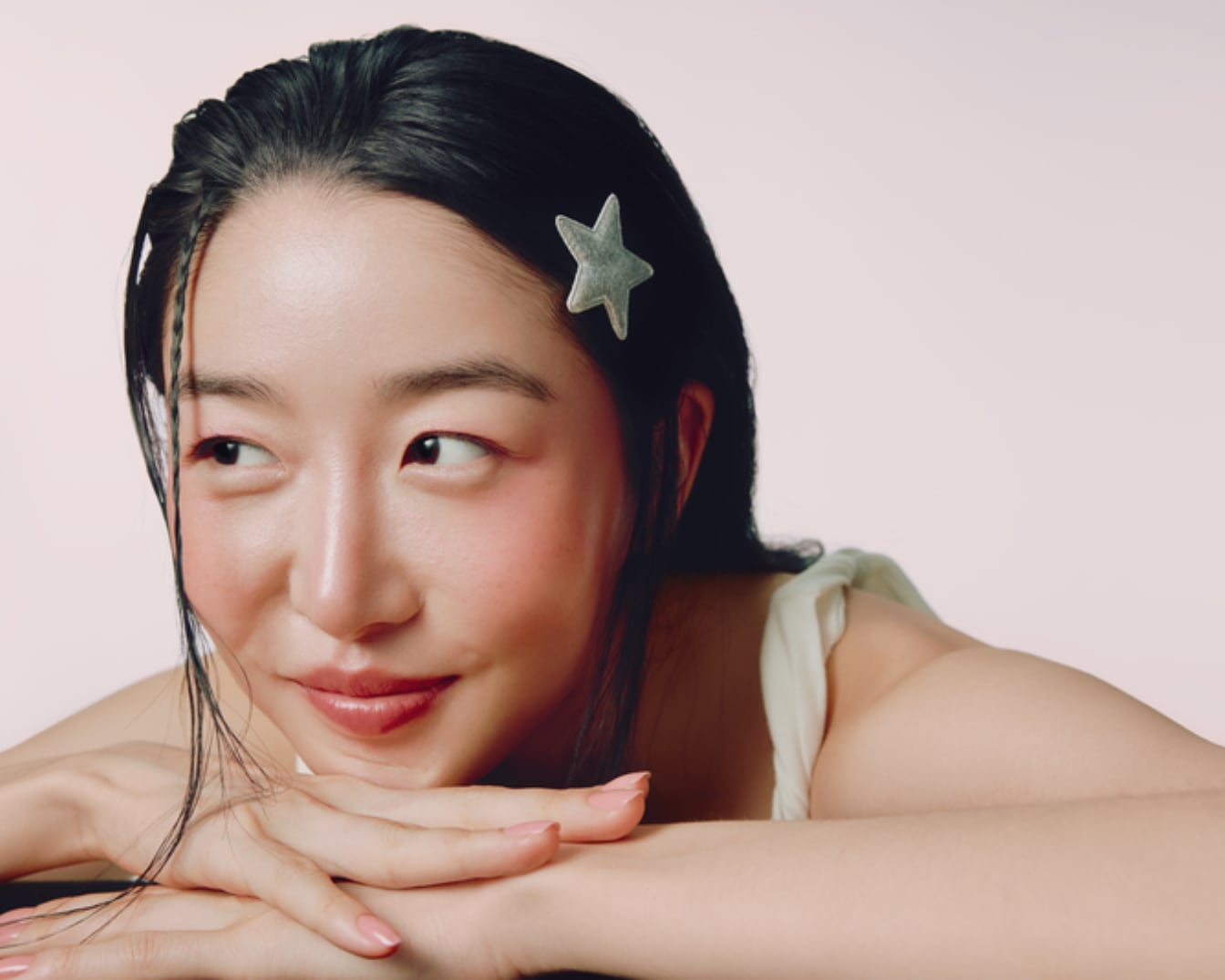 Une femme aux cheveux noirs et brillants, coiffée d'une barrette en forme d'étoile argentée, pose avec un maquillage naturel rehaussé d'un blush rosé sur les joues et d'un rouge à lèvres nude, les mains croisées sous le menton.