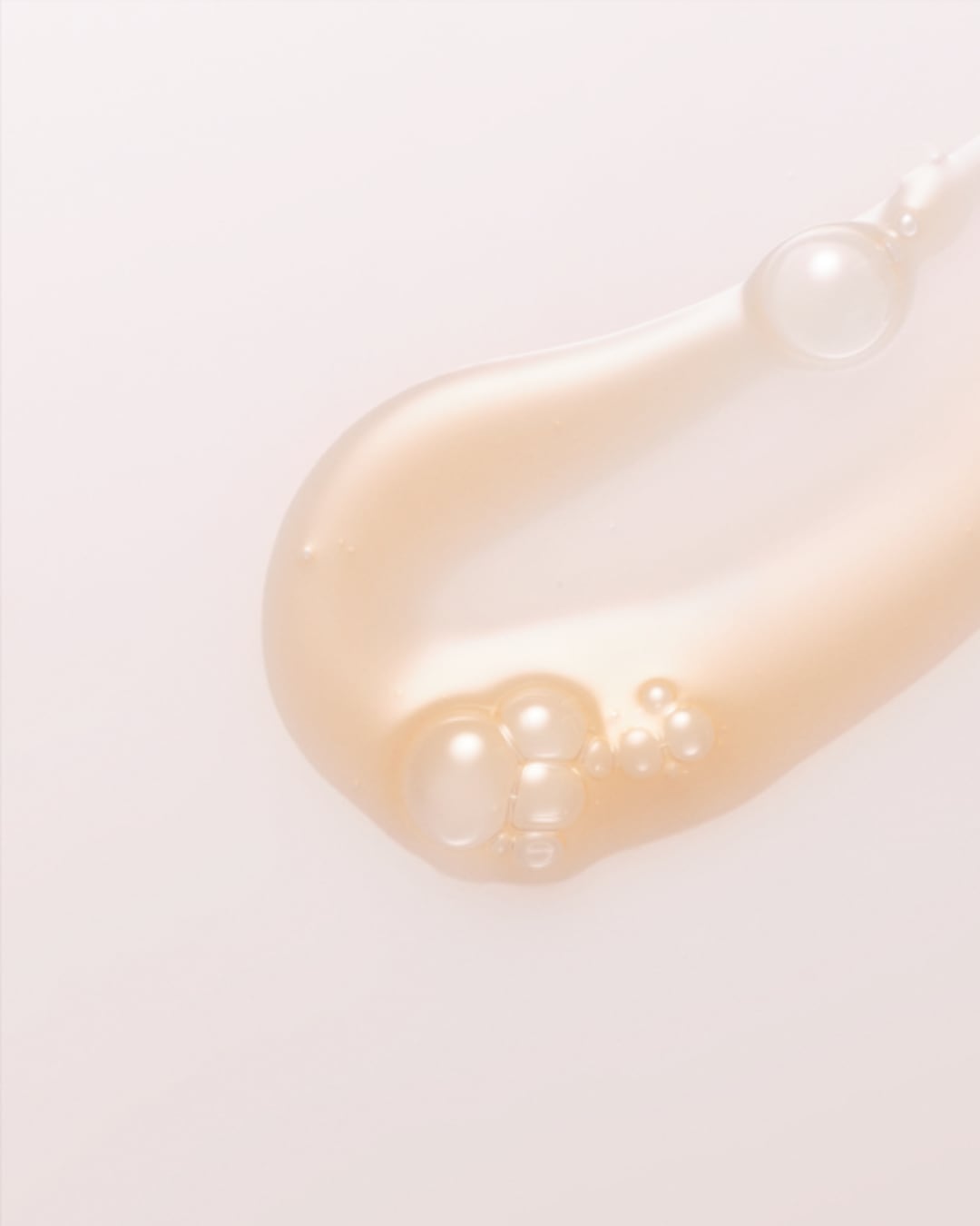 Une goutte de sérum doré avec des bulles d'air, étalée sur une surface claire, illustre la texture légère et la consistance du produit.