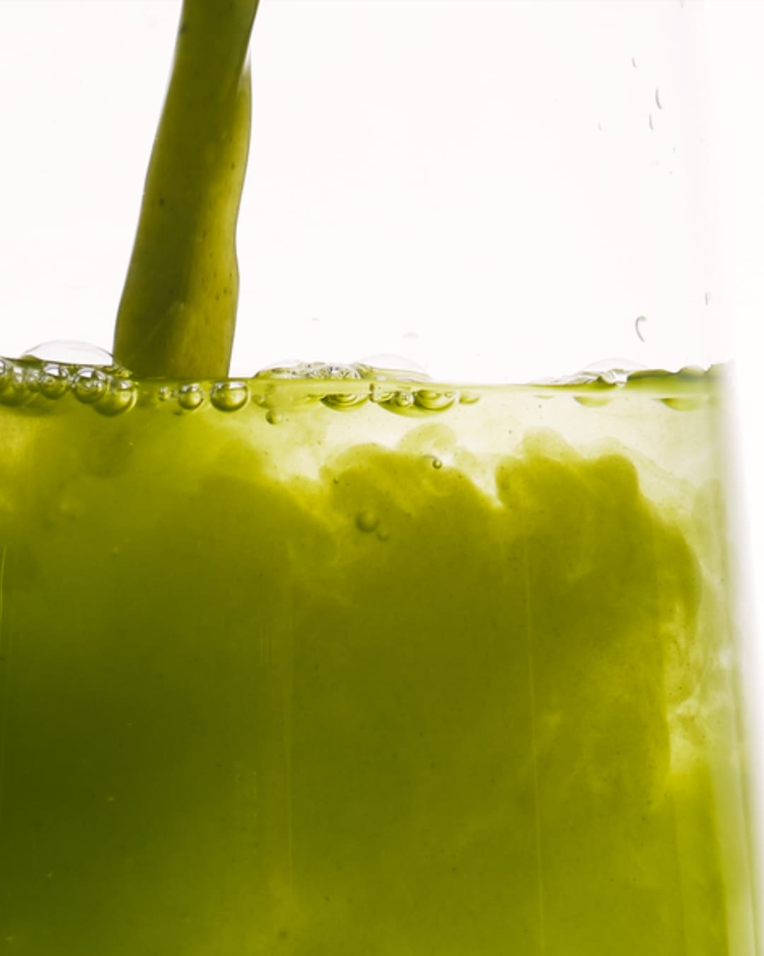 Un liquide vert vif est versé dans un verre transparent, créant des bulles et des tourbillons à la surface, suggérant une boisson rafraîchissante ou un ingrédient naturel.