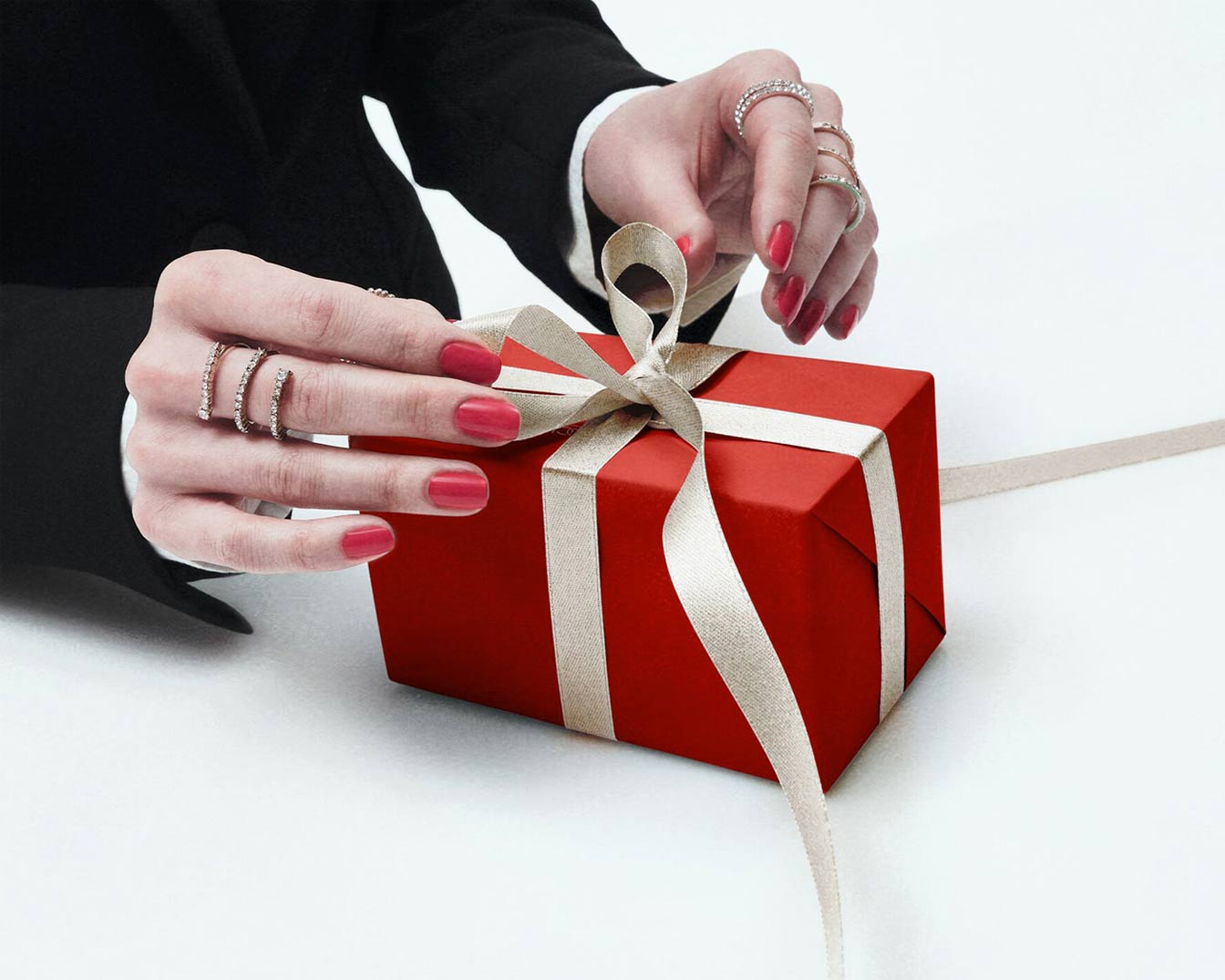 Une personne aux ongles rouges et aux bagues scintillantes noue un ruban beige sur une boîte cadeau rouge, prête à être offerte.