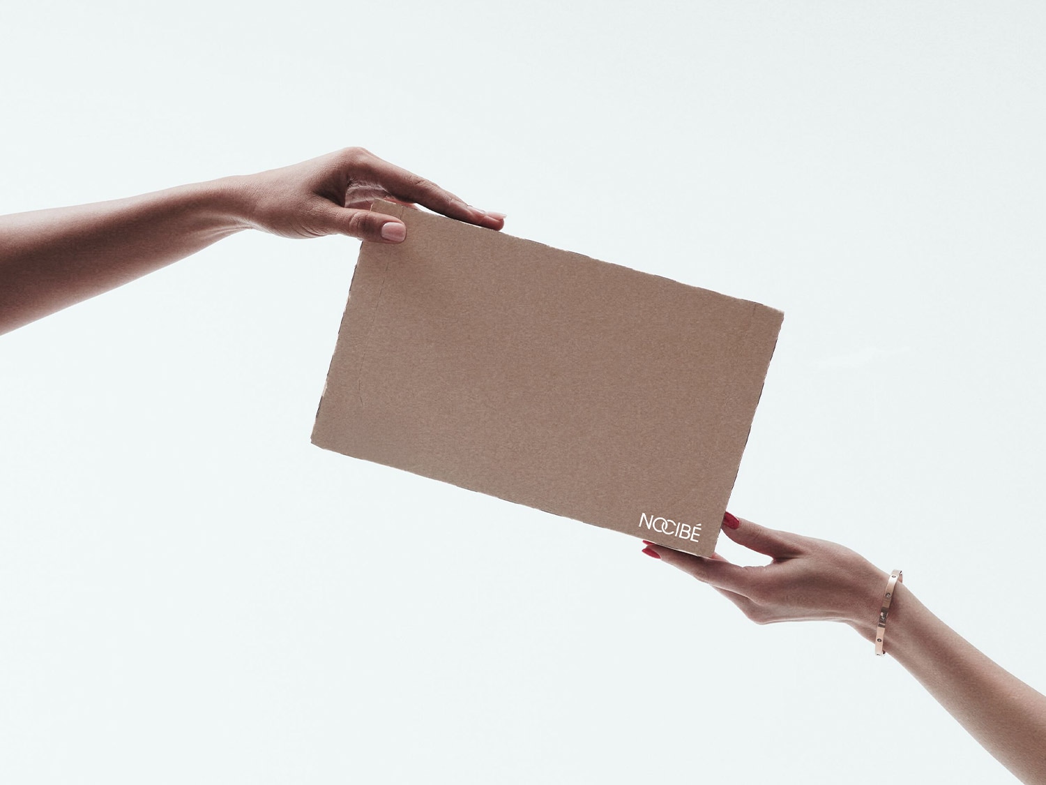 Deux mains tiennent une boîte en carton marron avec le logo blanc de Nocibé, l'une des mains porte un bracelet doré et du vernis à ongles rouge.