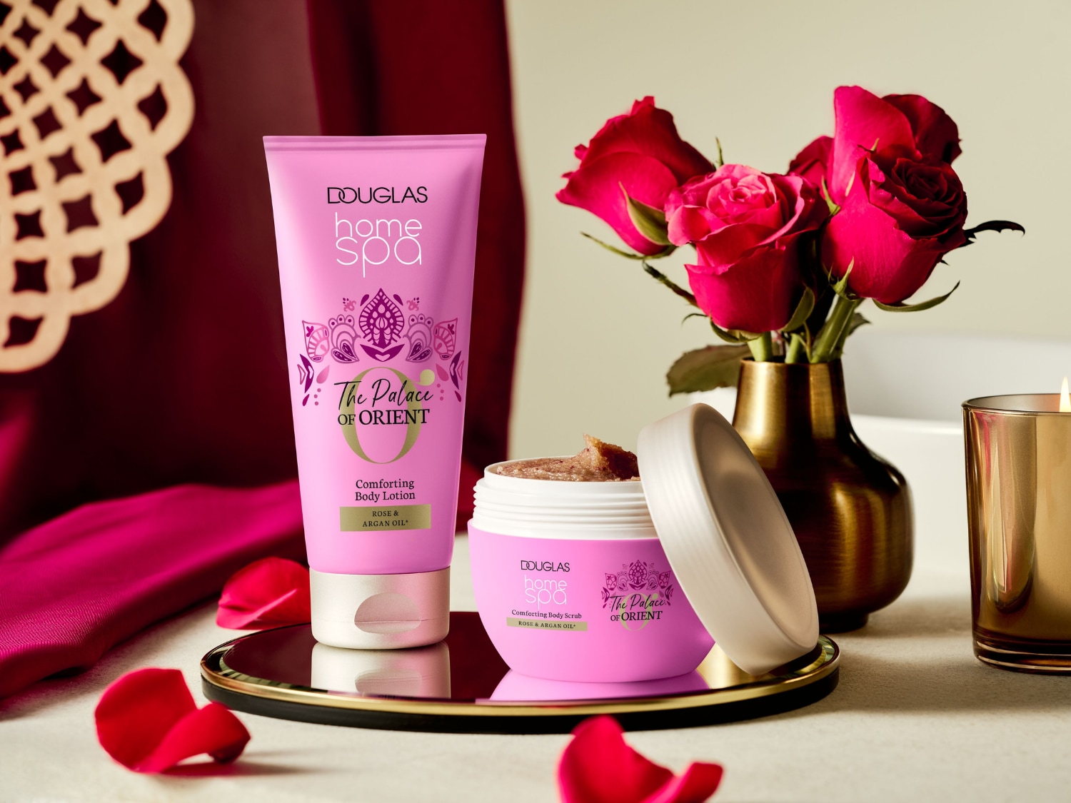 Różowy balsam do ciała Douglas Home Spa The Palace of Orient z różą i olejkiem arganowym, obok otwartego peelingu do ciała w słoiczku, na złotej tacy, z płatkami róż i świecą.
