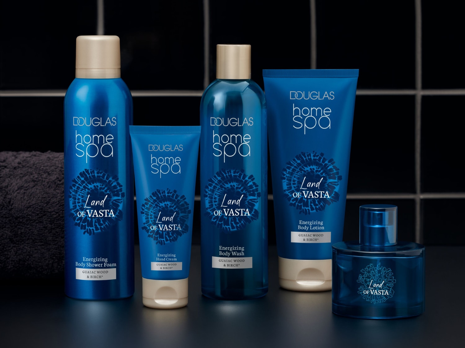 Kolekcja Douglas Home Spa Land of Vasta, w skład której wchodzą: pianka pod prysznic, krem do rąk, płyn do mycia ciała, balsam do ciała i woda toaletowa, wszystkie w eleganckich, niebieskich opakowaniach ze złotymi akcentami, z napisem "Land of Vasta" i wzorem przypominającym słońce.