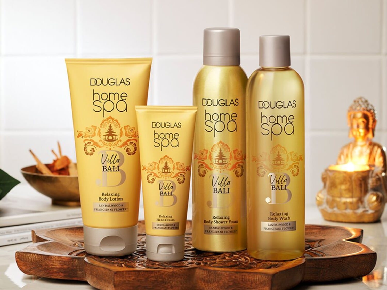 Zestaw Douglas Home Spa Villa Bali zawiera balsam do ciała, krem do rąk, piankę pod prysznic i płyn do mycia ciała, wszystkie w złotych opakowaniach z motywami kwiatowymi, stojące na drewnianej tacy obok figurki Buddy.