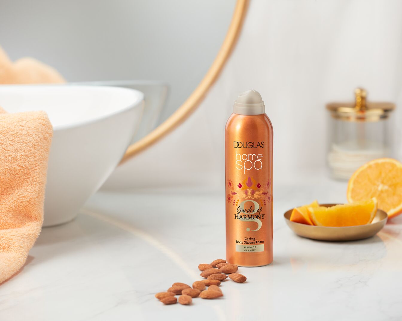 Douglas Home Spa Garden of Harmony to pielęgnująca pianka pod prysznic o zapachu migdałów i pomarańczy, w pomarańczowej butelce z ozdobnym wzorem, stojąca na marmurowej półce obok migdałów i kawałków pomarańczy.