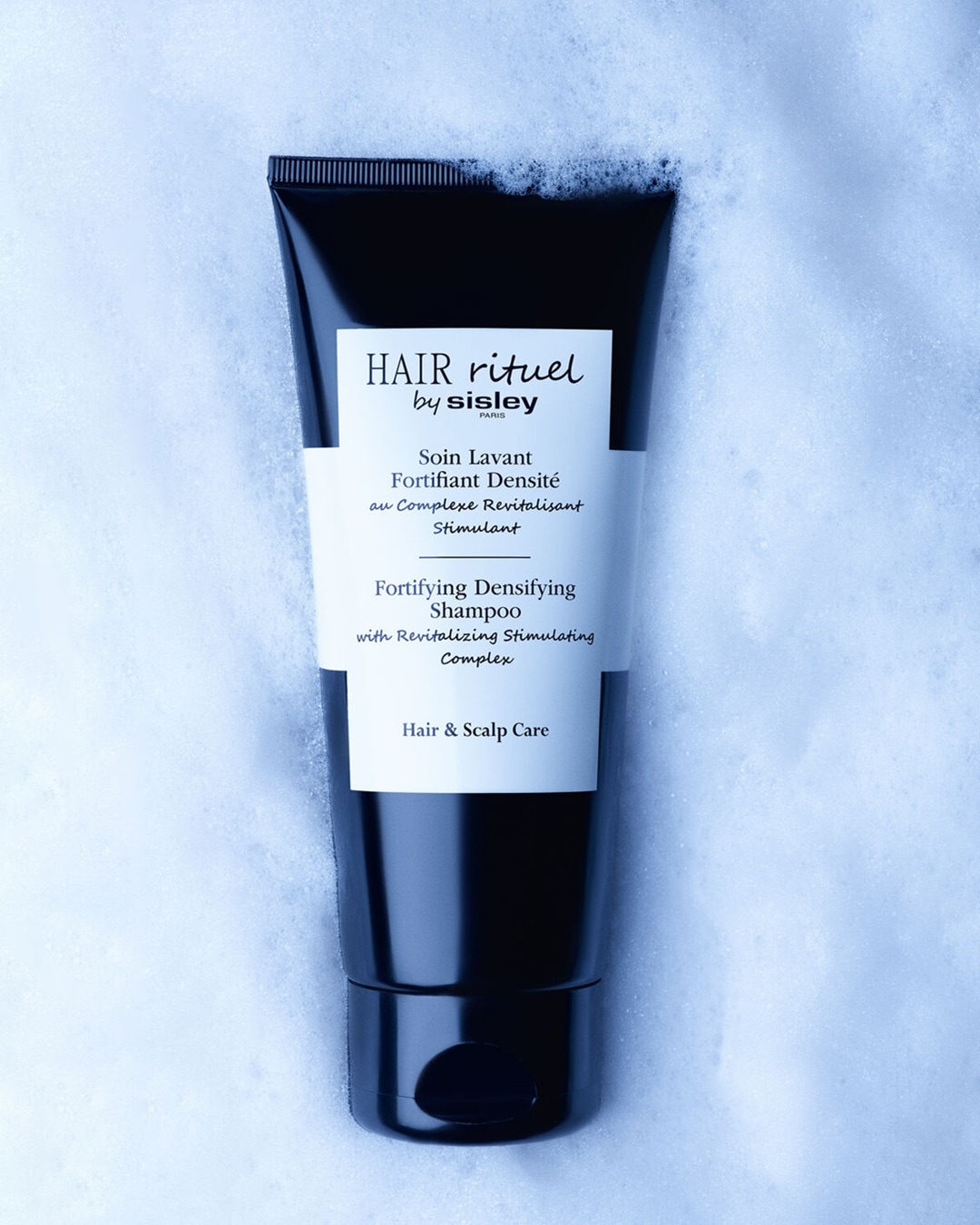 Szampon wzmacniający i zagęszczający Hair Rituel by Sisley w czarnej tubce leży na białej, spienionej powierzchni, z widocznym napisem "Hair & Scalp Care".