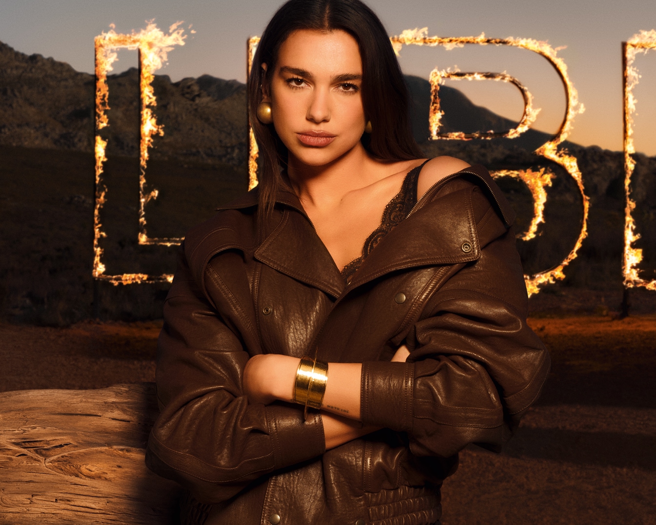 Dua Lipa poseert zelfverzekerd in een bruine leren jas met een kanten top, haar armen gekruist, tegen een achtergrond van bergen en vlammende letters die "LIBRE" spellen.