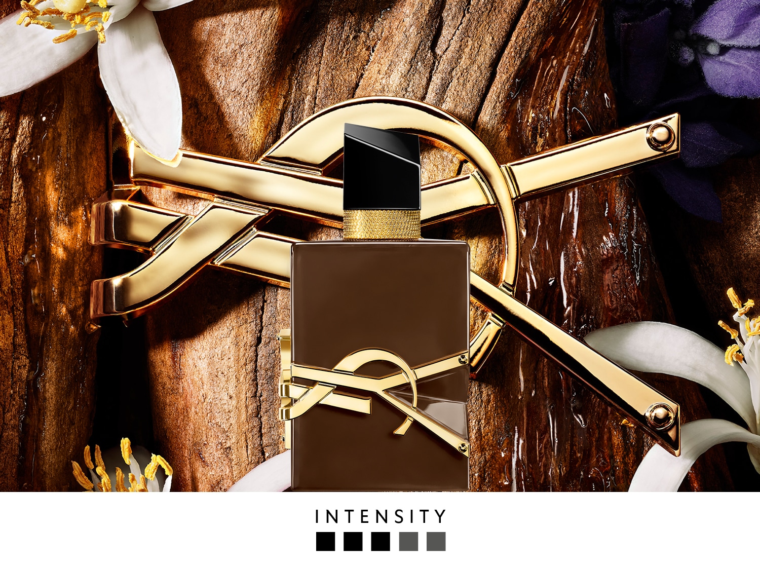 Een bruine parfumfles met een gouden YSL-logo en een zwarte dop staat op een houten oppervlak, omringd door witte en paarse bloemblaadjes, met het woord "INTENSITY" eronder.