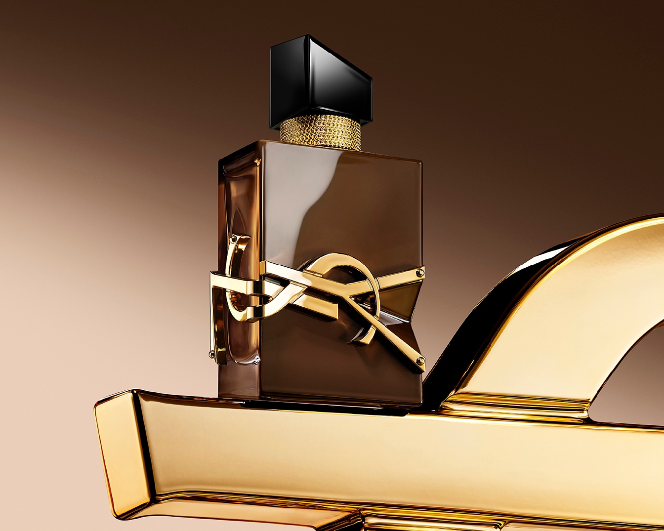 Een elegante parfumfles van Yves Saint Laurent, met een donkerbruine vloeistof en een glanzend gouden YSL-logo, staat op een gouden, sculpturale basis tegen een zachtbruine achtergrond.