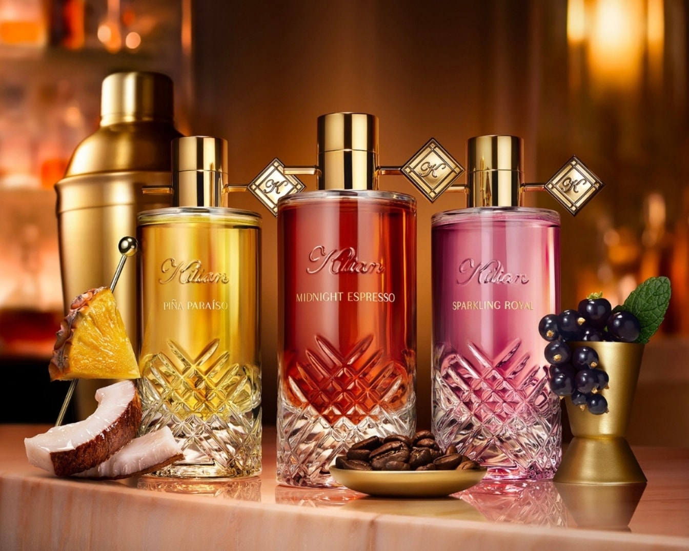 Drie Kilian Paris parfumflessen, Piña Paraíso, Midnight Espresso en Sparkling Royal, staan op een bar met cocktailingrediënten zoals ananas, kokosnoot, koffiebonen en bessen.