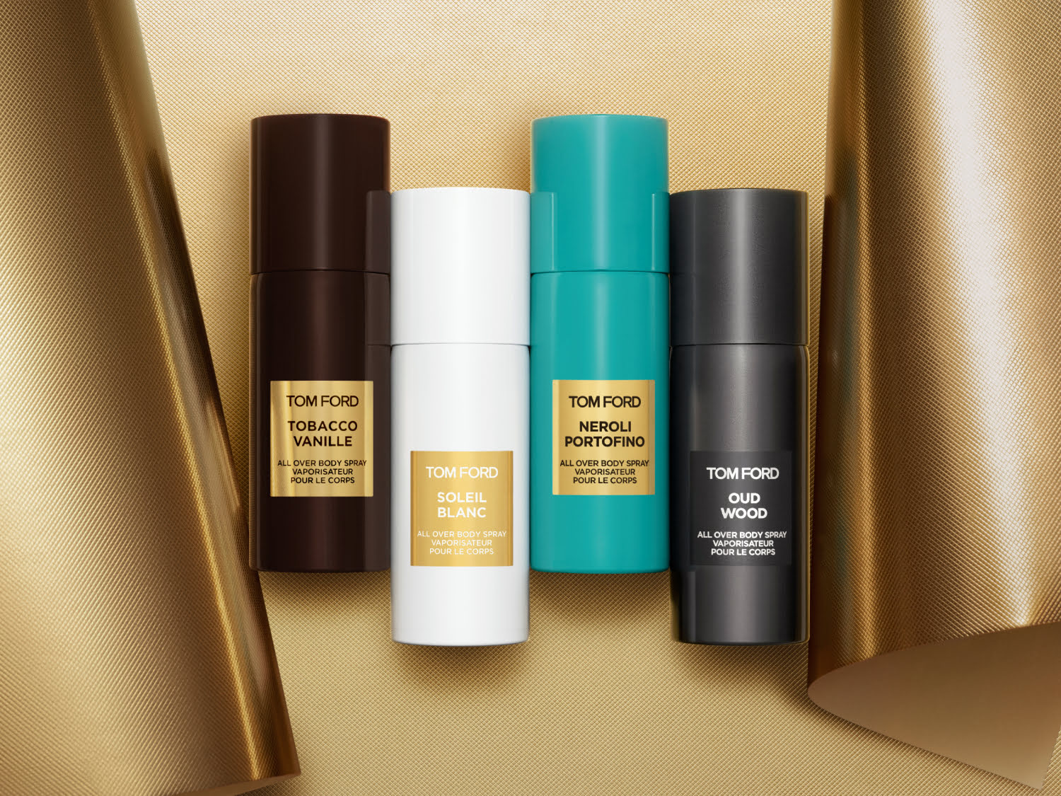 Keturios TOM FORD kūno purškiklių pakuotės, rudos, baltos, turkio ir juodos spalvos, išdėstytos ant auksinio gofruoto paviršiaus, su užrašais „Tobacco Vanille“, „Soleil Blanc“, „Neroli Portofino“ ir „Oud Wood“.