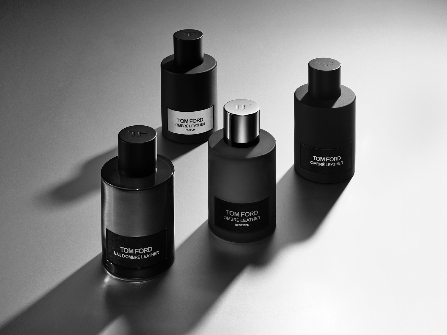 Keturi Tom Ford Ombré Leather kvepalų buteliukai, išdėstyti ant pilko paviršiaus, sukuria elegantišką ir rafinuotą kompoziciją.