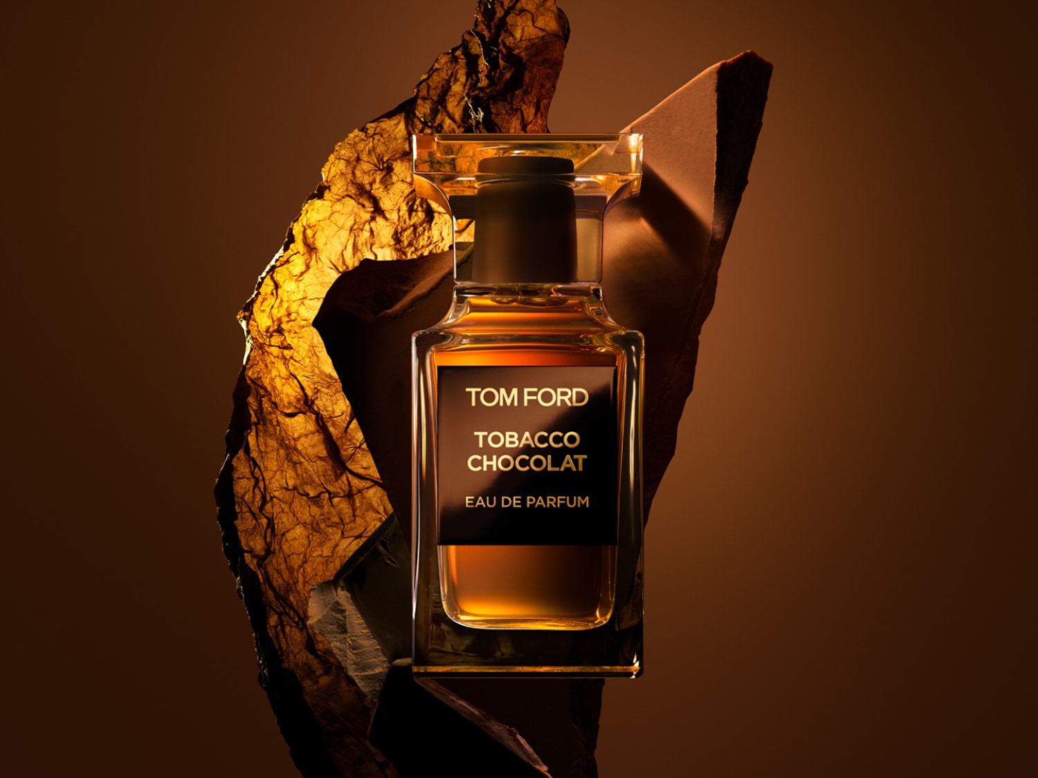 Tom Ford Tobacco Chocolat kvepalai, auksinės spalvos skysčiu, stovi ant rudos uolos, o fone – ruda siena.