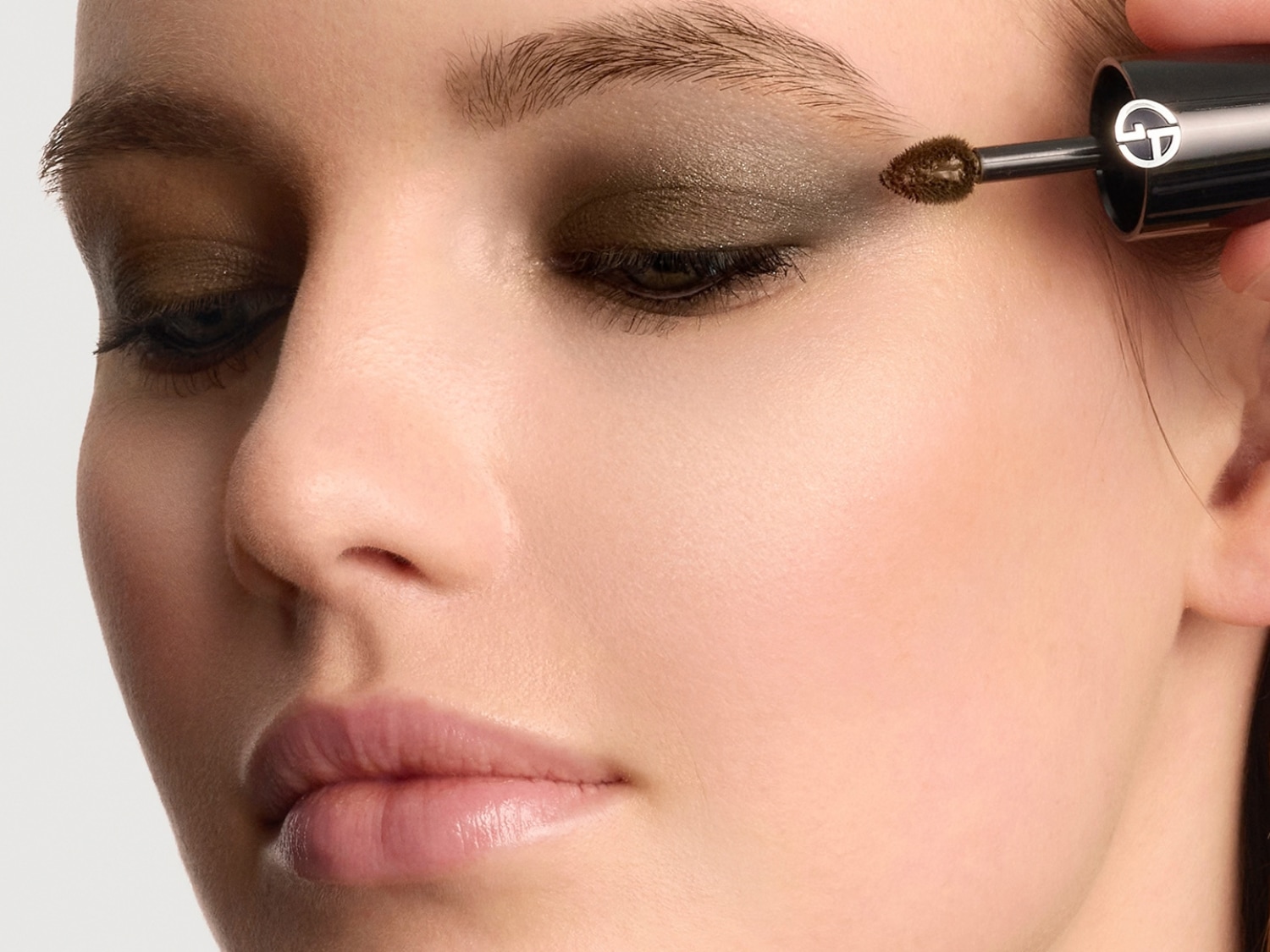 Žena si nanáší tmavě zelené oční stíny na víčko pomocí aplikátoru s logem Giorgio Armani, což zvýrazňuje její líčení.