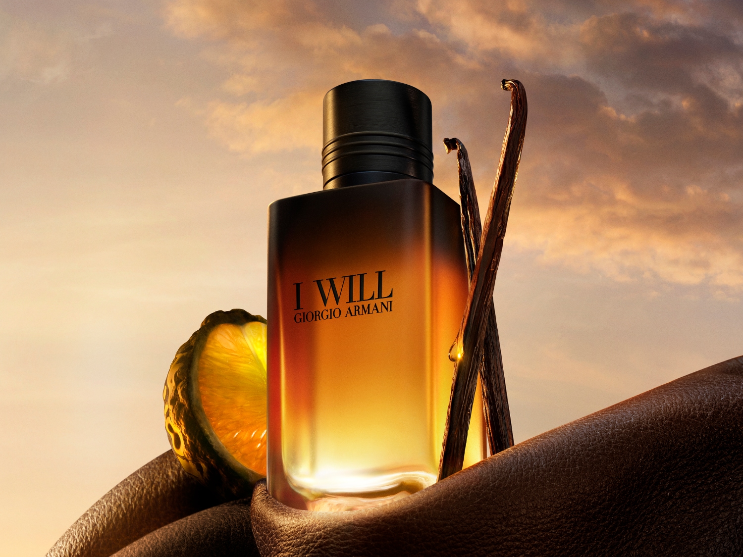 Az "I WILL GIORGIO ARMANI" feliratú parfümös üveg narancssárga és barna árnyalatokban pompázik, mellette egy félbevágott citrusfélét és vaníliarudakat láthatunk, melyek egy barna bőr textúrájú anyagon fekszenek, a háttérben naplementés égbolt látszik.