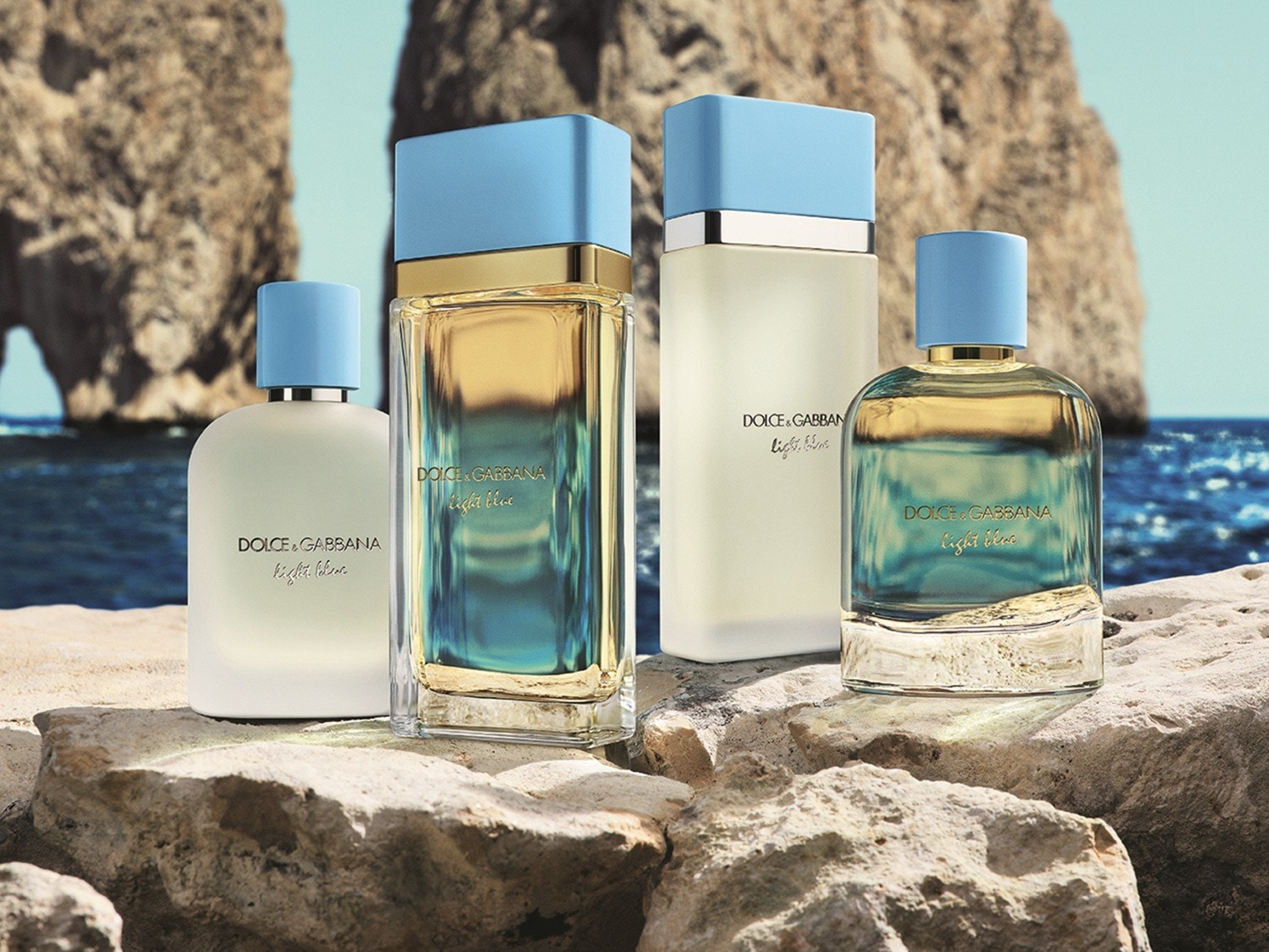 Vier Dolce & Gabbana Light Blue parfums, twee in helder glas met goudkleurige vloeistof en twee in matglas met een lichtere vloeistof, staan op rotsen aan de kust met de zee en kliffen op de achtergrond.
