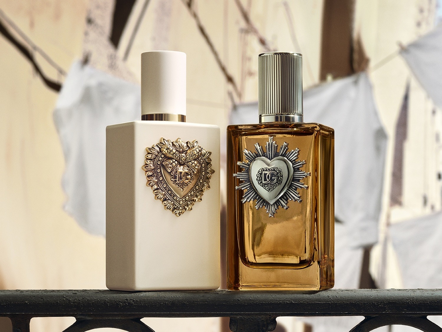 Twee Dolce & Gabbana Devotion parfums staan op een donkergrijze reling, met de linker fles in mat crème en de rechter in glanzend amberkleurig glas, beide versierd met een hartvormig medaillon.