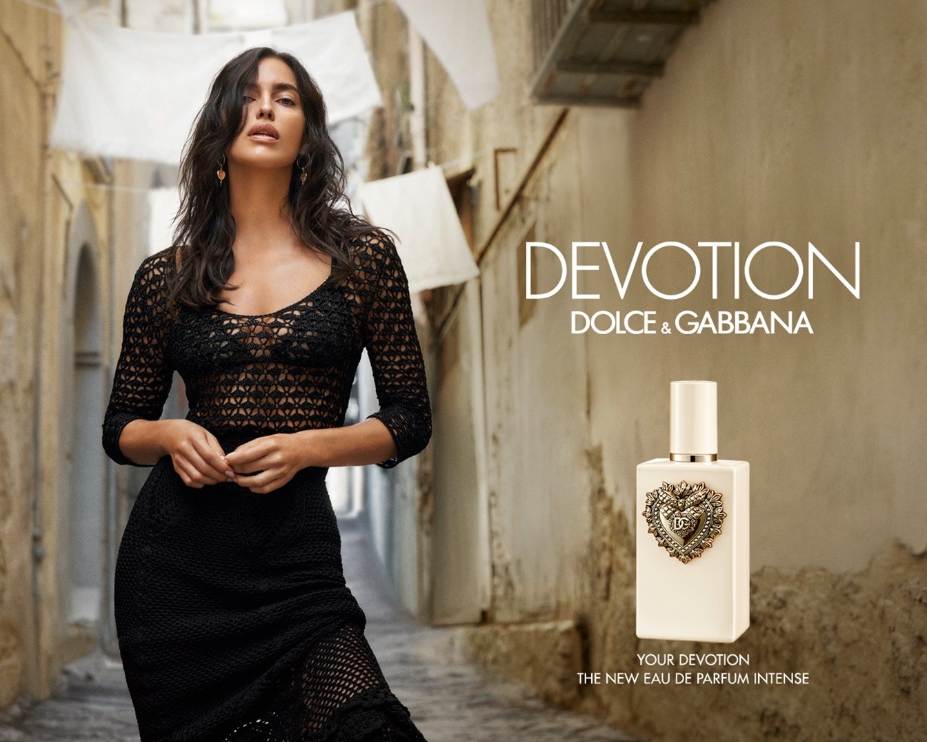 Une femme aux cheveux foncés, vêtue d'une robe en maille noire, pose dans une ruelle étroite, avec le parfum Devotion de Dolce & Gabbana à ses côtés.