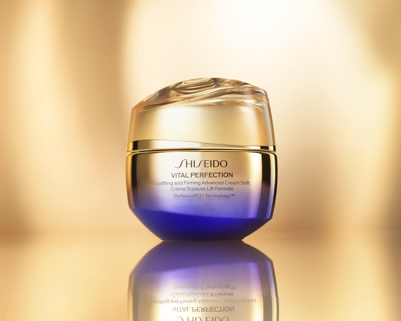 Shiseido Vital Perfection Uplifting and Firming Advanced Cream Soft, un vasetto di crema viso con coperchio dorato e base viola sfumata, è posizionato su una superficie riflettente dorata.