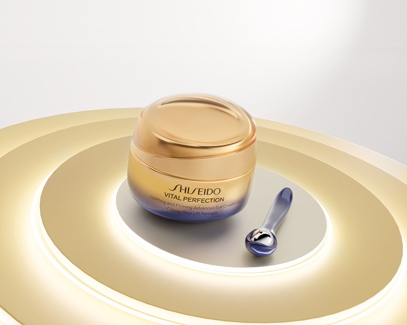 Shiseido Vital Perfection Uplifting and Firming Advanced Eye Cream in un lussuoso barattolo dorato e blu, con un applicatore massaggiante, è posizionata su una base illuminata.