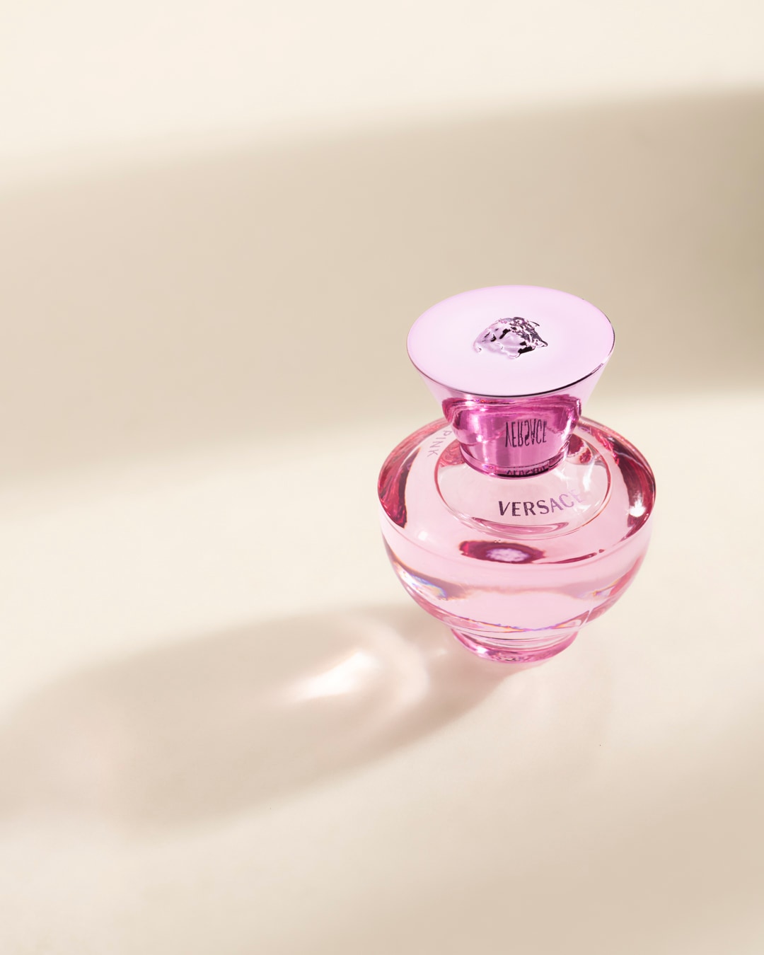 Le parfum Versace Bright Crystal est présenté dans un flacon en verre rose avec un bouchon rose orné du logo de la marque, posé sur une surface claire avec des ombres douces.