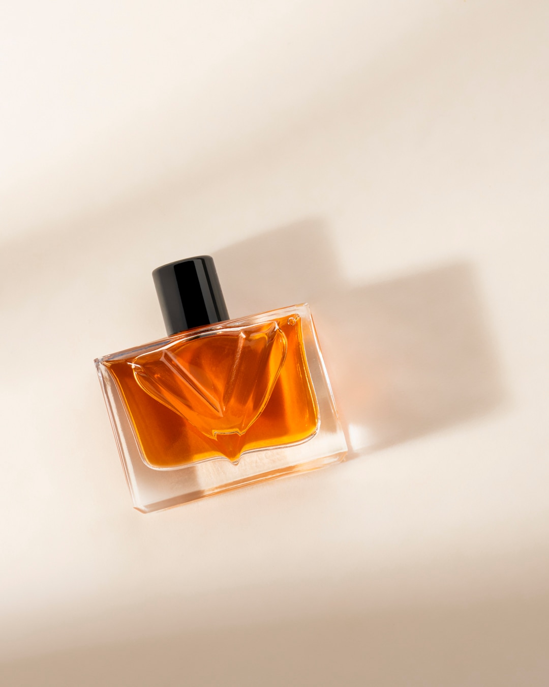 Un flacon de parfum en verre transparent avec un liquide ambré et un bouchon noir, posé en diagonale sur une surface beige clair avec des ombres douces.