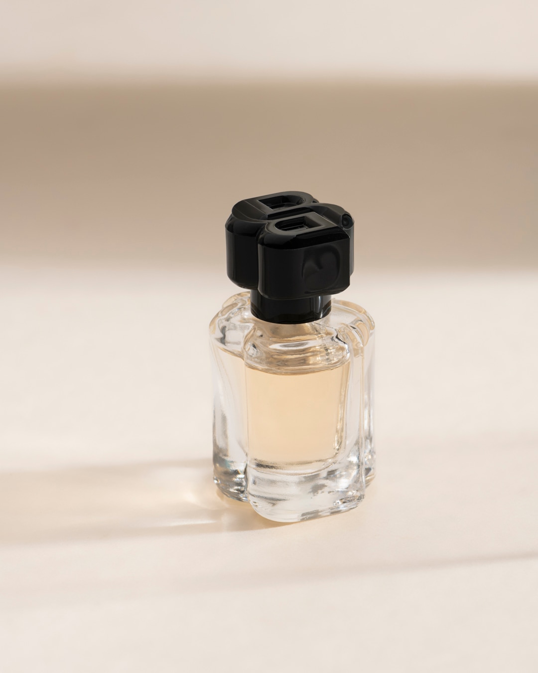 Un flacon de parfum en verre transparent avec un liquide ambré et un bouchon noir orné d'un motif en forme de « B » stylisé, posé sur une surface claire avec des ombres douces.