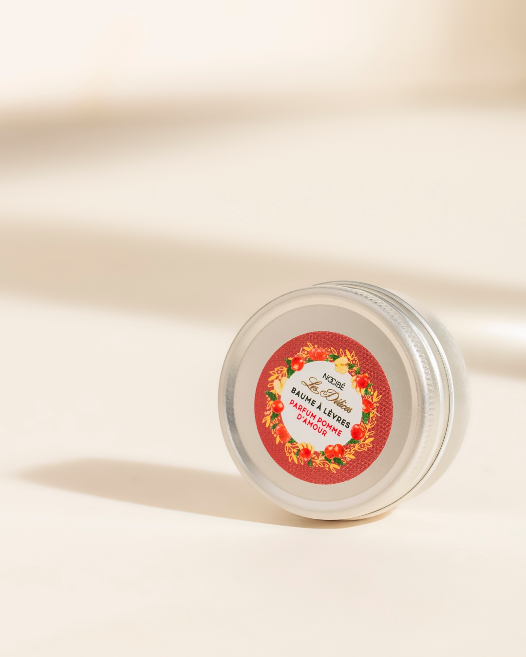 Un baume à lèvres Nocibé Les Délices Parfum Pomme d'Amour dans un pot en métal argenté, avec une étiquette rouge ornée de pommes et de feuilles, posé sur une surface claire.