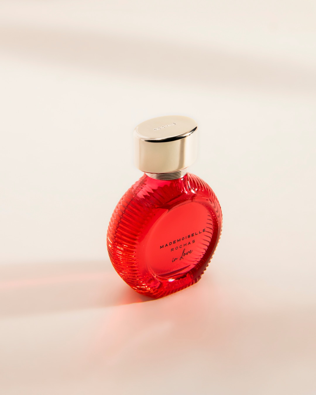 Le parfum Mademoiselle Rochas In Love est présenté dans un flacon rouge vif avec un bouchon argenté, posé sur une surface claire.