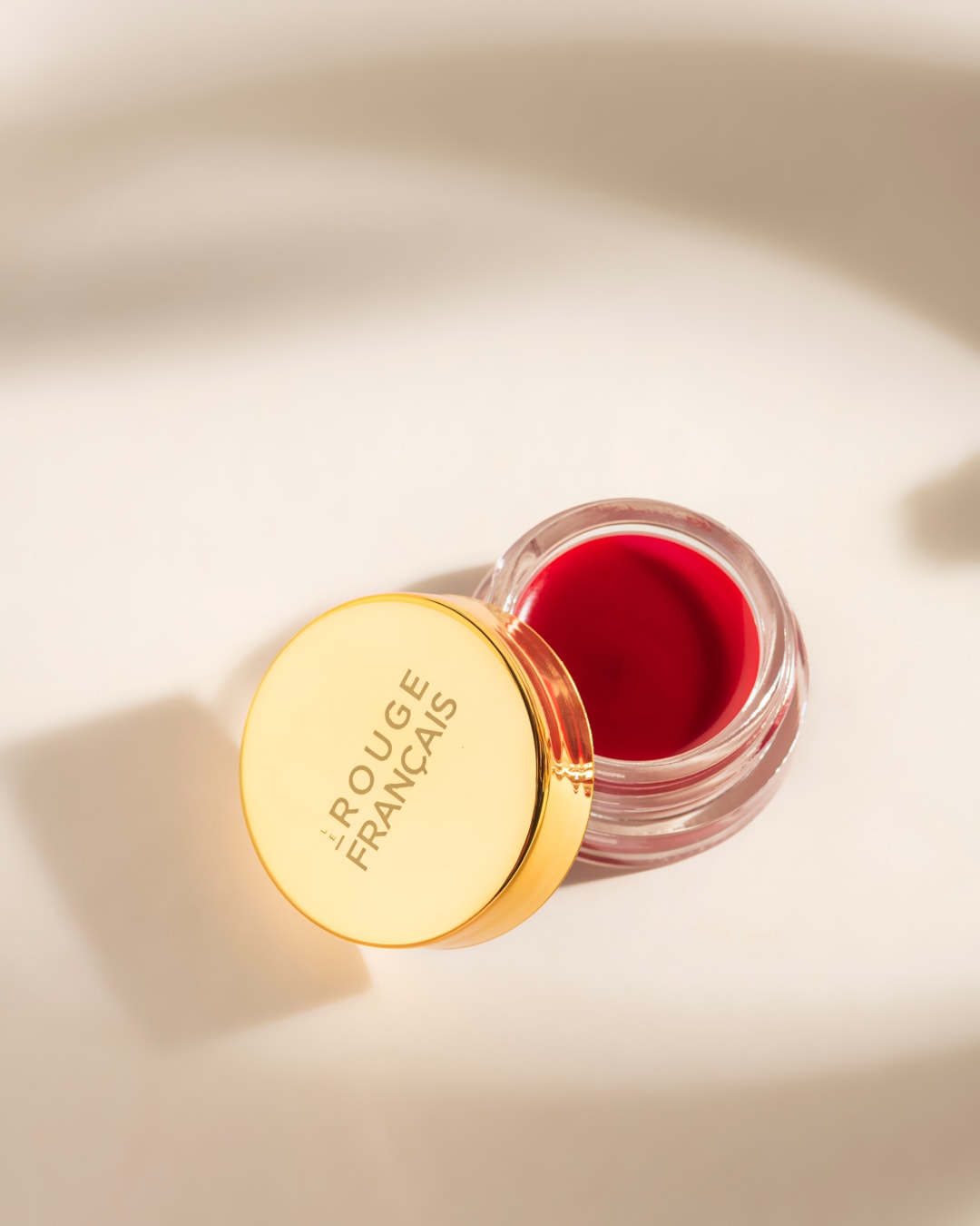 Un pot de baume à lèvres rouge vif de Le Rouge Français, avec un couvercle doré portant le nom de la marque, est posé sur une surface claire avec des ombres douces.