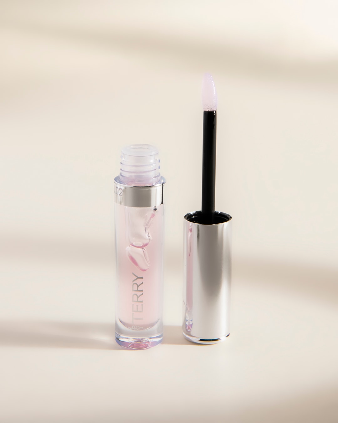 Le brillant à lèvres By Terry est présenté dans un flacon transparent avec un applicateur à baguette, révélant une teinte rose pâle et une texture brillante.