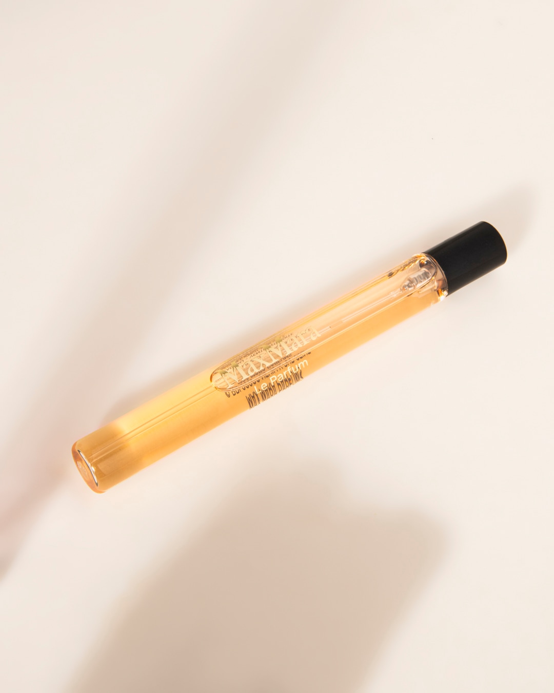Un flacon de parfum Max Mara Le Parfum, de forme cylindrique et allongé, est posé sur une surface blanche, son liquide ambré et son bouchon noir contrastant avec l'étiquette dorée.