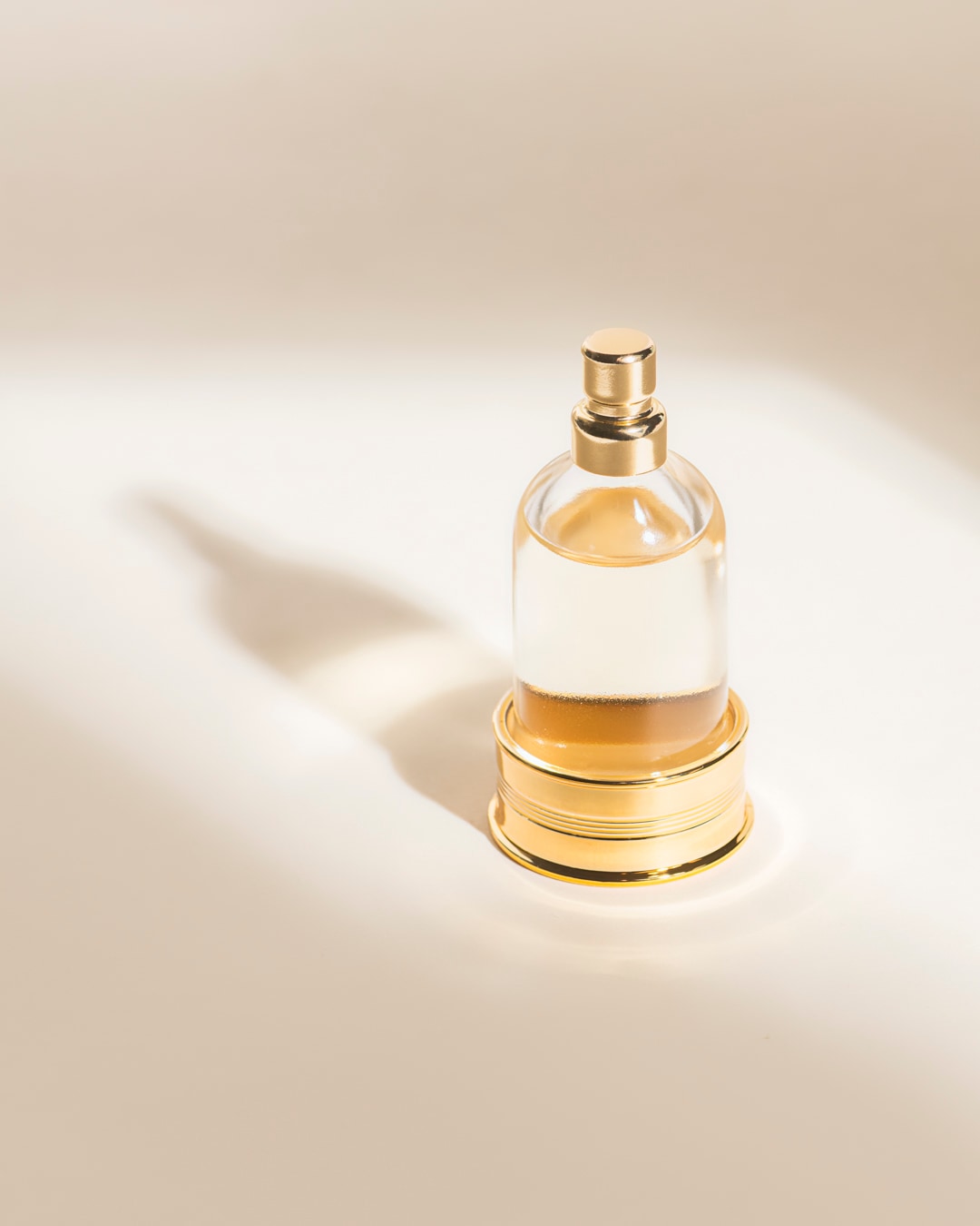 Un flacon de parfum en verre transparent avec un bouchon et une base dorés, rempli d'un liquide ambré, est posé sur une surface claire avec des ombres douces.