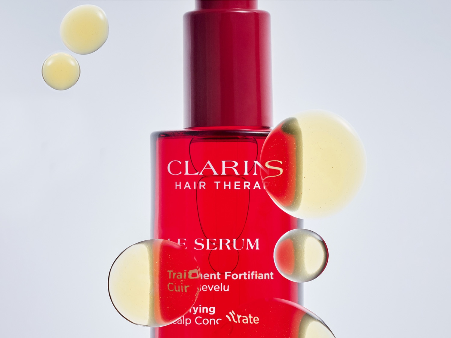 Serul Clarins Hair Therapy, într-o sticlă roșie vibrantă, este prezentat cu picături aurii de ulei care plutesc în jurul său, sugerând o formulă bogată și hrănitoare pentru păr.