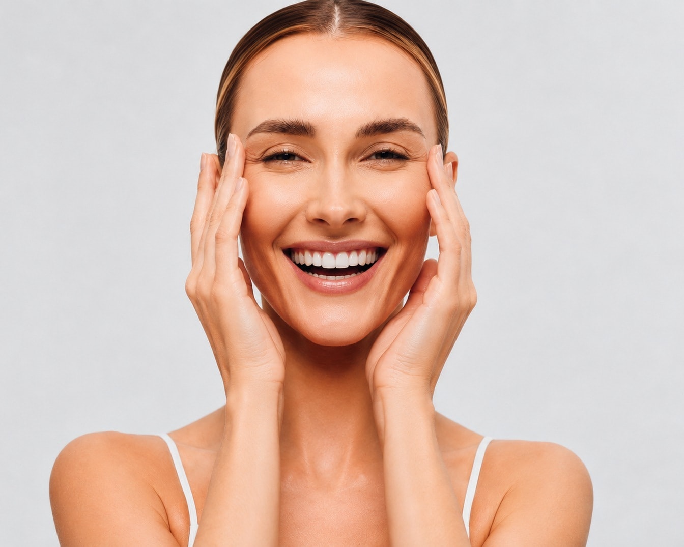 Una donna sorridente con i capelli raccolti all'indietro, indossa una canotta bianca e tiene le mani ai lati del viso, mettendo in risalto la sua pelle luminosa e le rughe d'espressione intorno agli occhi.