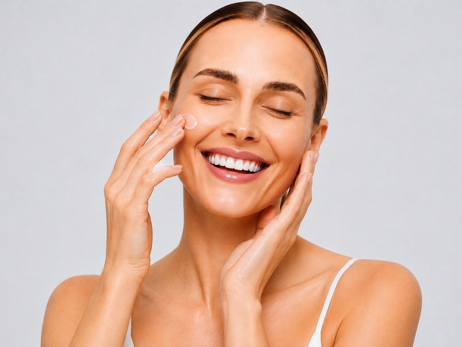 Una donna sorridente con gli occhi chiusi applica una crema idratante sul viso, con una piccola quantità di prodotto visibile sulla guancia, per una pelle luminosa e idratata.