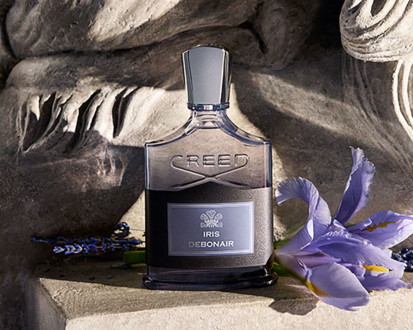 Descoperiți parfumul Creed Iris Debonair, o sticlă elegantă cu etichetă gri, așezată pe o suprafață de piatră, alături de o floare de iris mov și lavandă, evocând o aromă sofisticată.