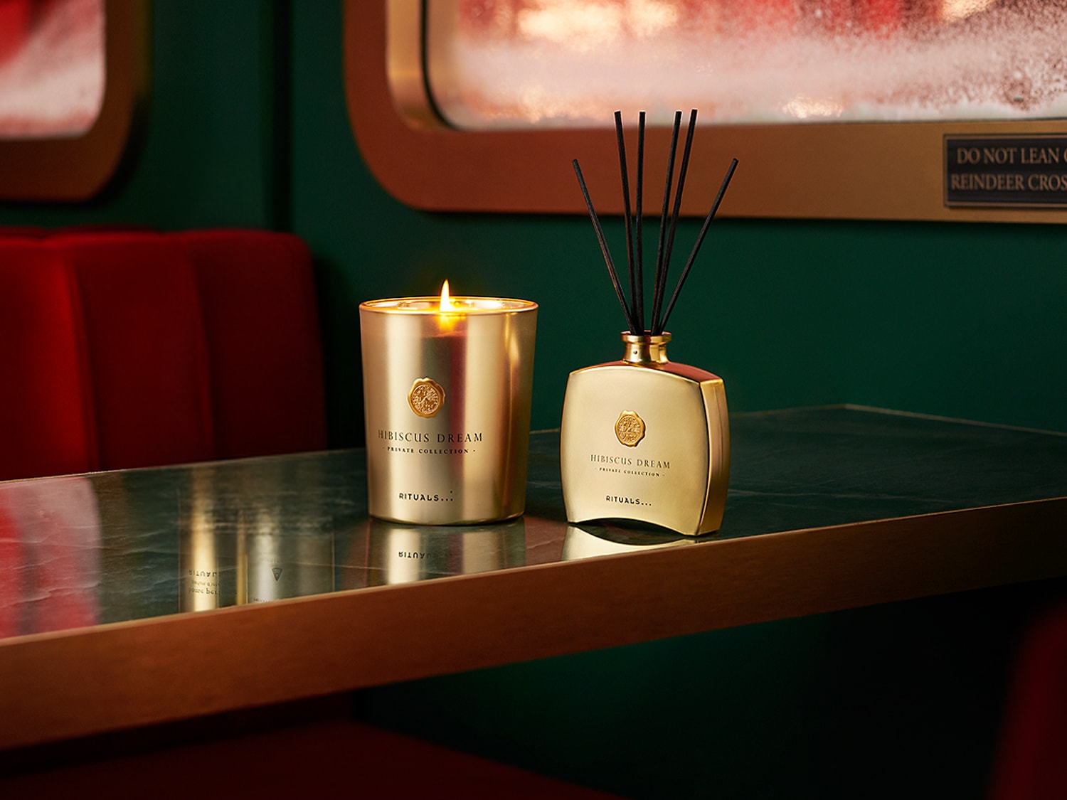 O lumânare aurie aprinsă și un difuzor de parfum cu bețișoare negre, ambele cu eticheta "Hibiscus Dream Private Collection Rituals", sunt așezate pe o masă lucioasă, într-un cadru festiv cu banchete roșii și pereți verzi.