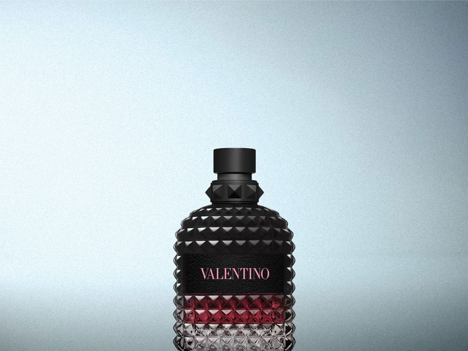 Le parfum Valentino Uomo Born in Roma Coral Fantasy est présenté dans une bouteille en verre noir texturé avec des détails cloutés, un capuchon noir et un liquide rouge-rose visible à travers le verre transparent à la base, avec le nom "VALENTINO" en rose sur une étiquette noire.