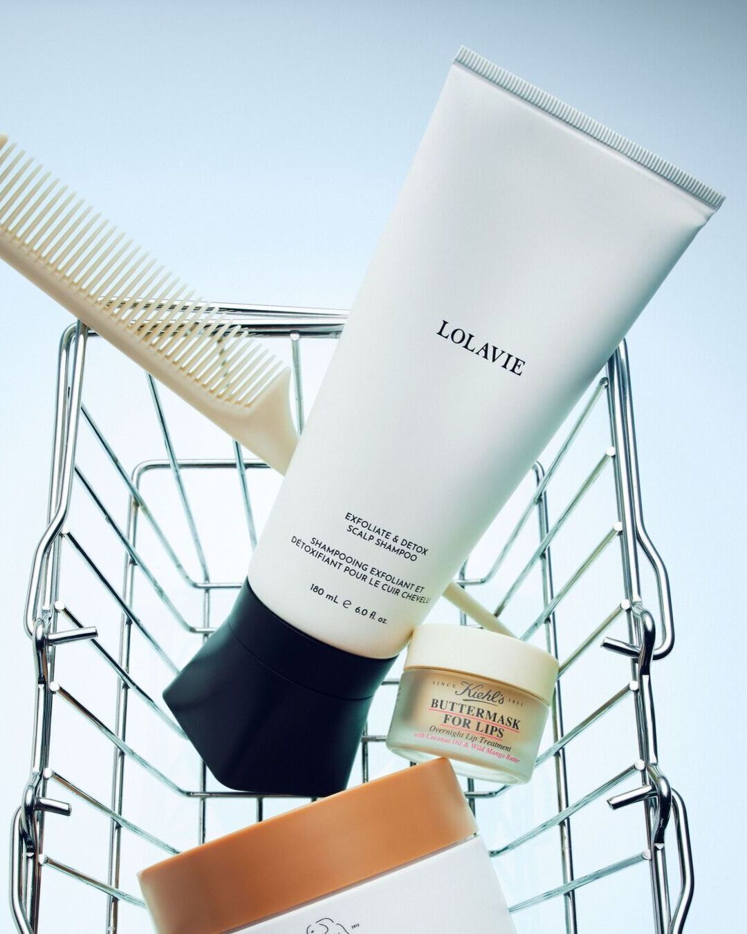 Šampūnas „LolaVie Exfoliate & Detox Scalp Shampoo“ ir „Kiehl's Buttermask for Lips Overnight Lip Treatment“ yra metaliniame krepšelyje su šukomis, skirtomis plaukų priežiūrai.