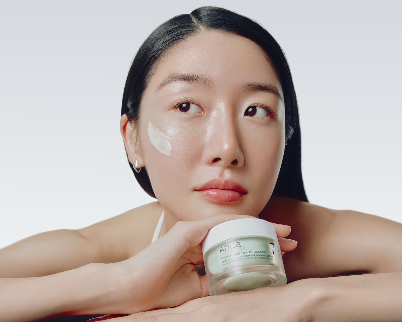 Una donna con i capelli scuri e lucidi tiene in mano un vasetto di crema Anua Heartleaf 70 + Ceramide, con una piccola quantità di prodotto applicata sulla sua guancia.