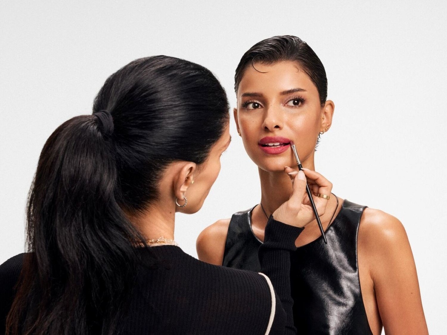 Una truccatrice applica un rossetto rosa intenso sulle labbra di una modella con un pennello, mentre la modella guarda in alto a destra con un'espressione serena.