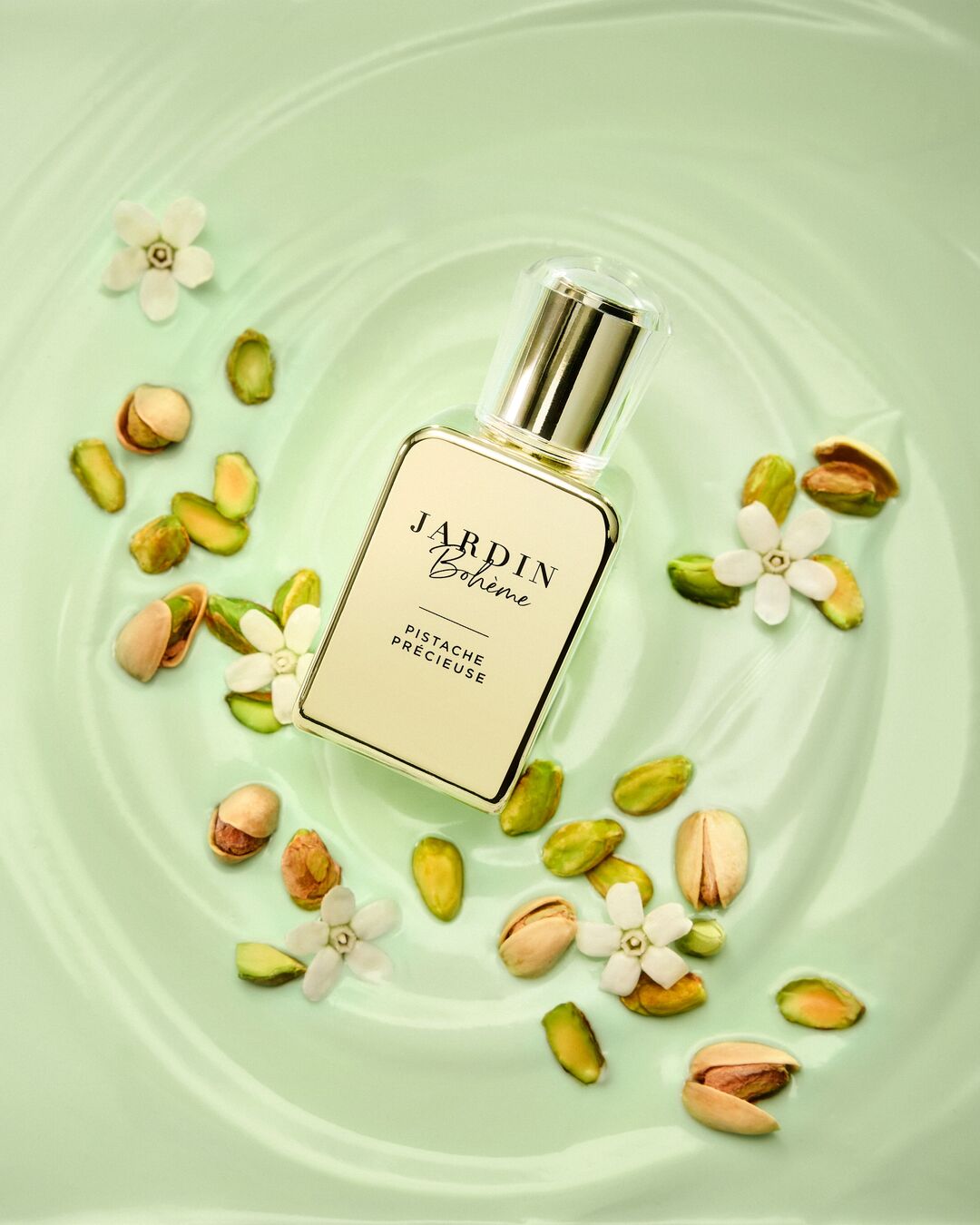 Le parfum Jardin Bohème Pistache Précieuse est présenté dans un flacon rectangulaire en verre transparent avec un bouchon doré, posé sur une surface vert clair, entouré de pistaches décortiquées et de petites fleurs blanches.